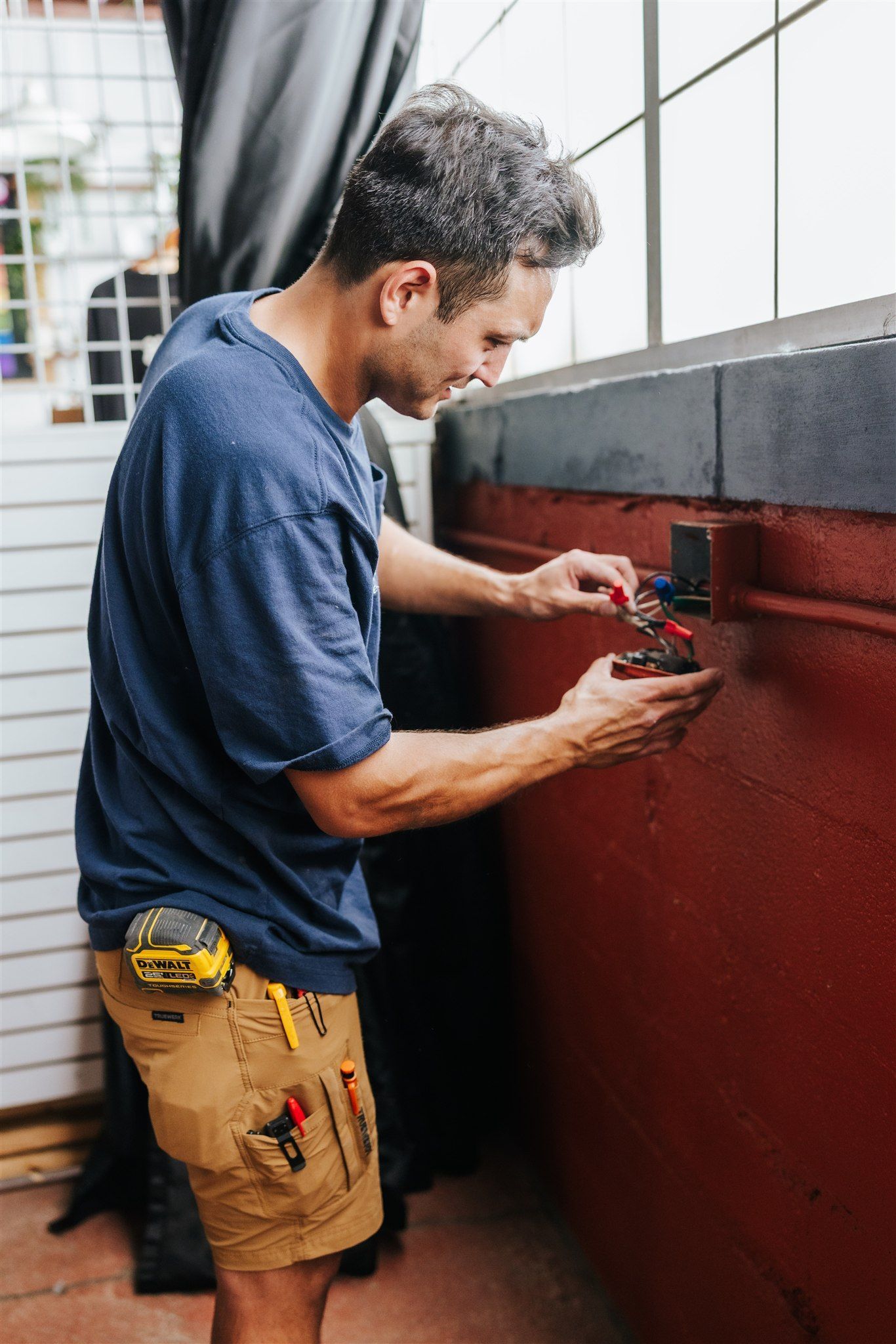 A person in a blue t-shirt and tan work shorts installs electrical wiring into a wall outlet on a red surface.