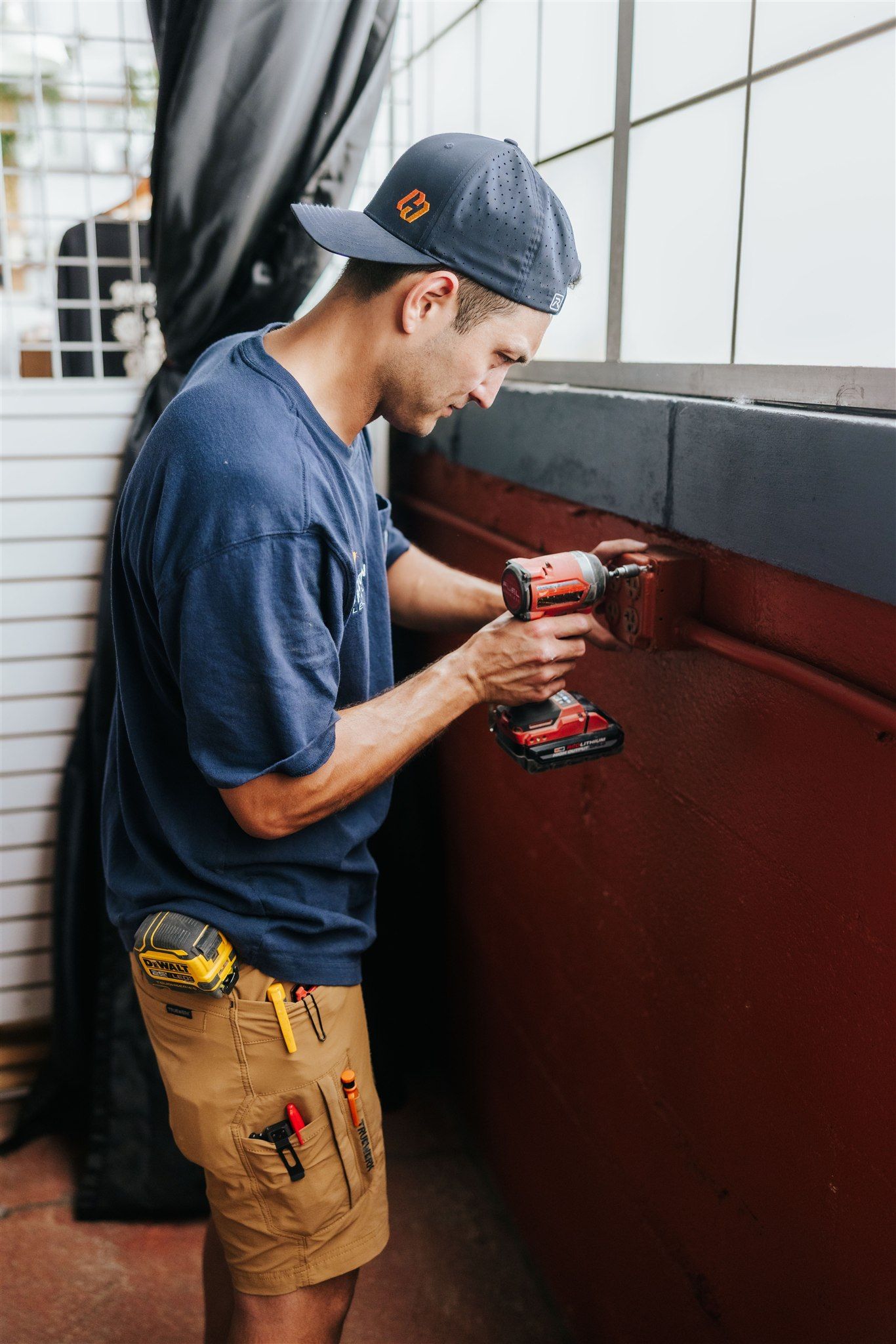 A worker in a blue shirt and khaki work shorts uses a red power drill on a dark red wall.