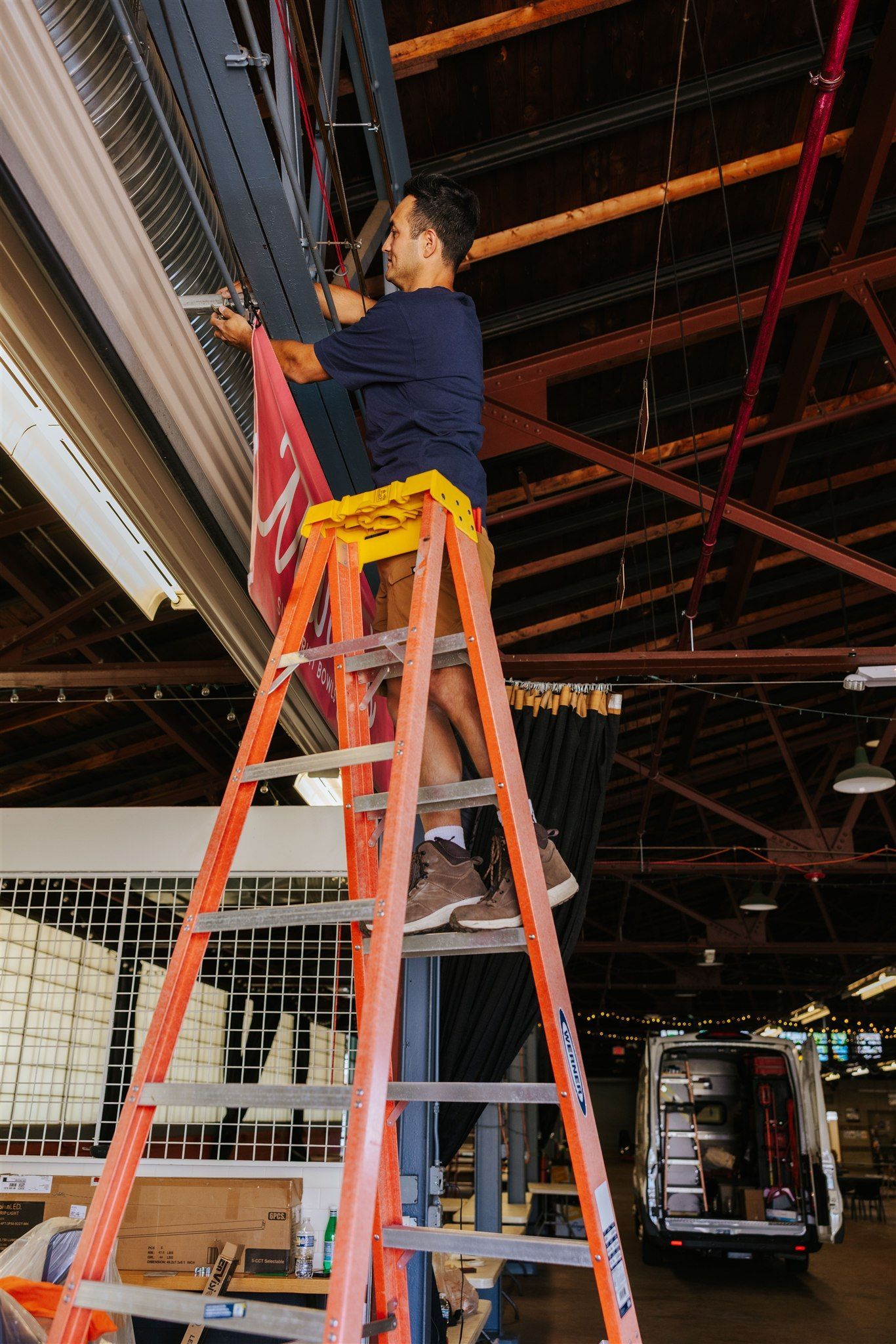 A person in a blue shirt standing on a tall orange ladder, attaching a red banner to a warehouse ceiling structure.