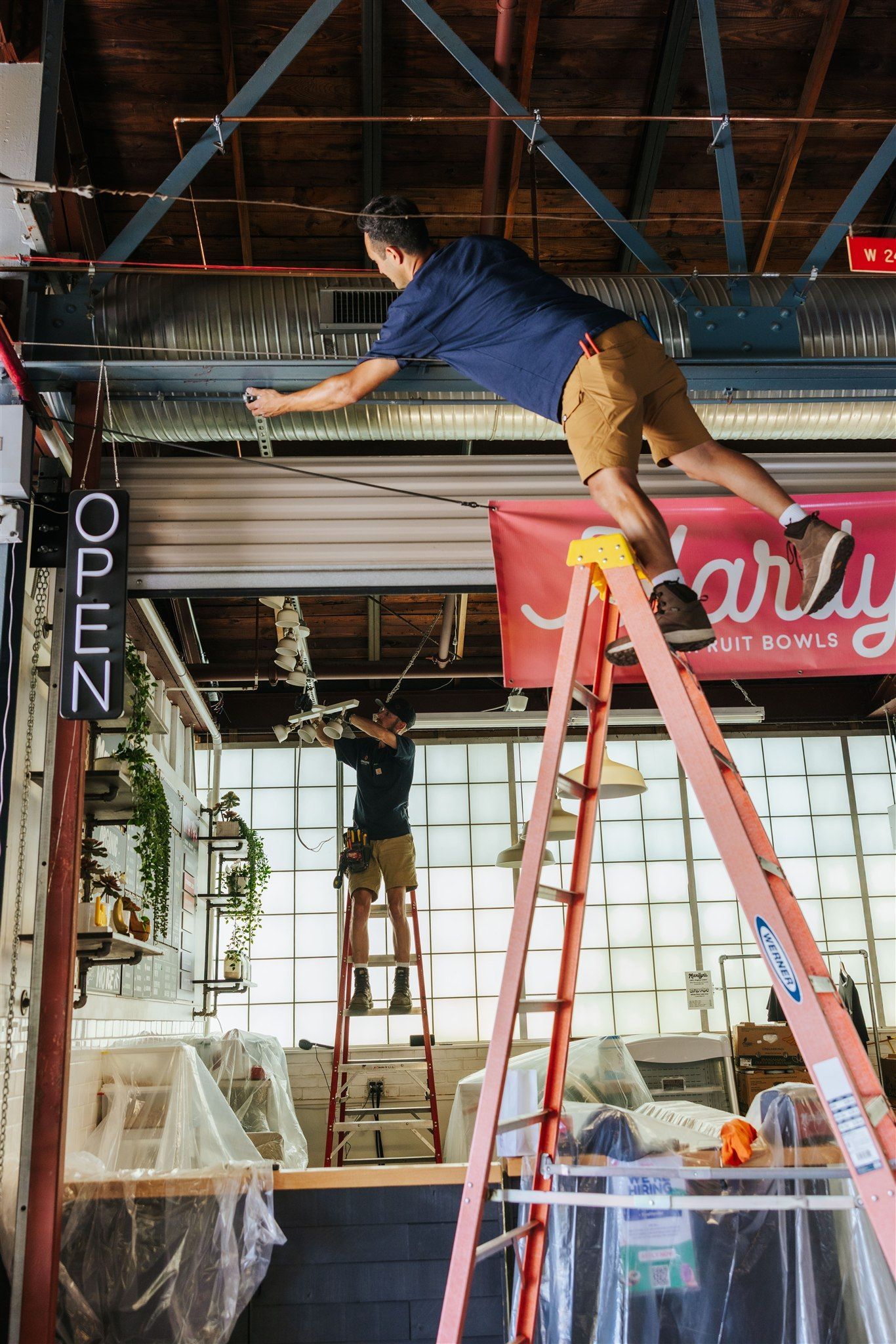 Two people on stepladders perform maintenance work on the exposed ceiling and lighting of a commercial space.