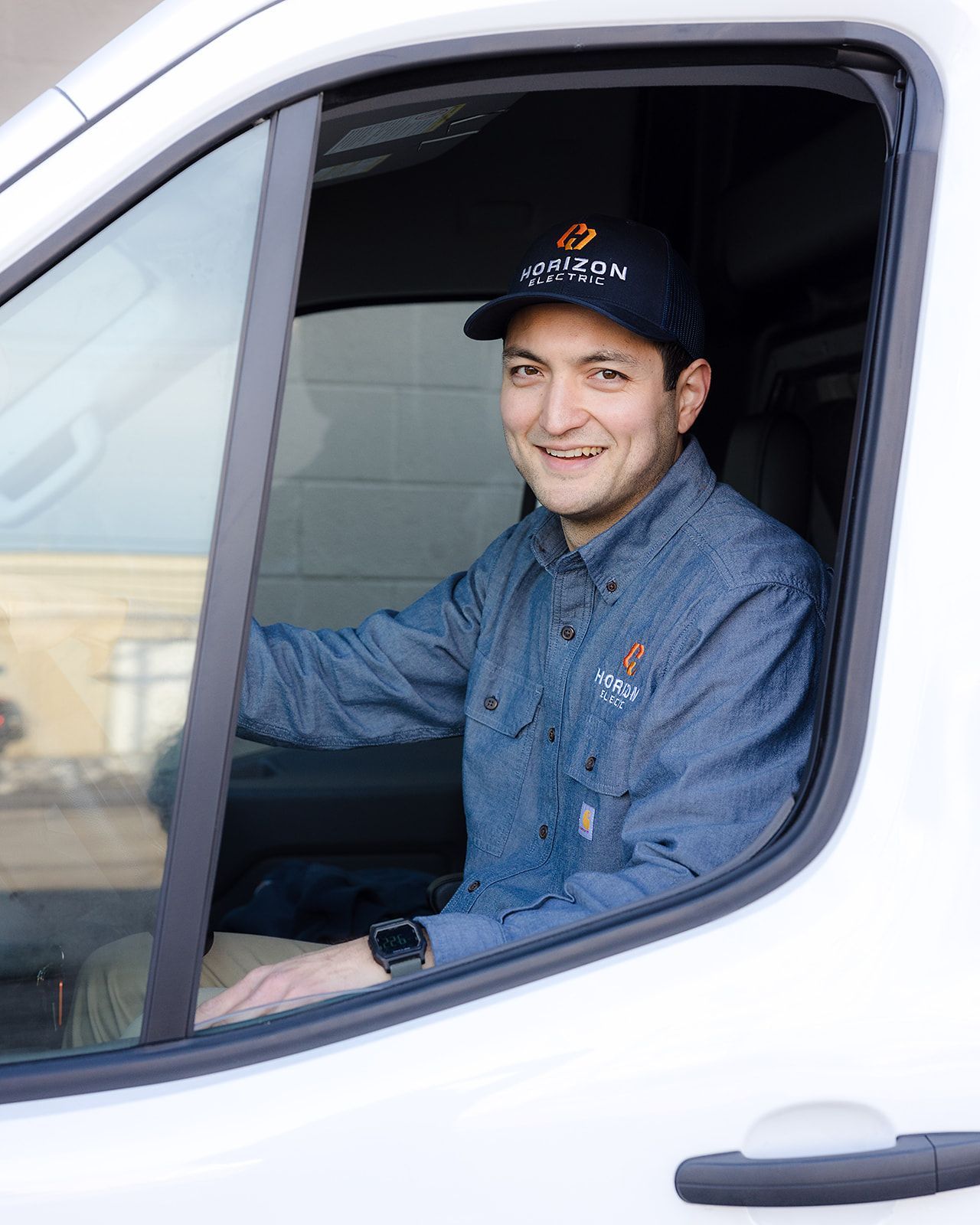 A person wearing a branded cap and blue shirt smiles while seated in the driver's seat of a white van.
