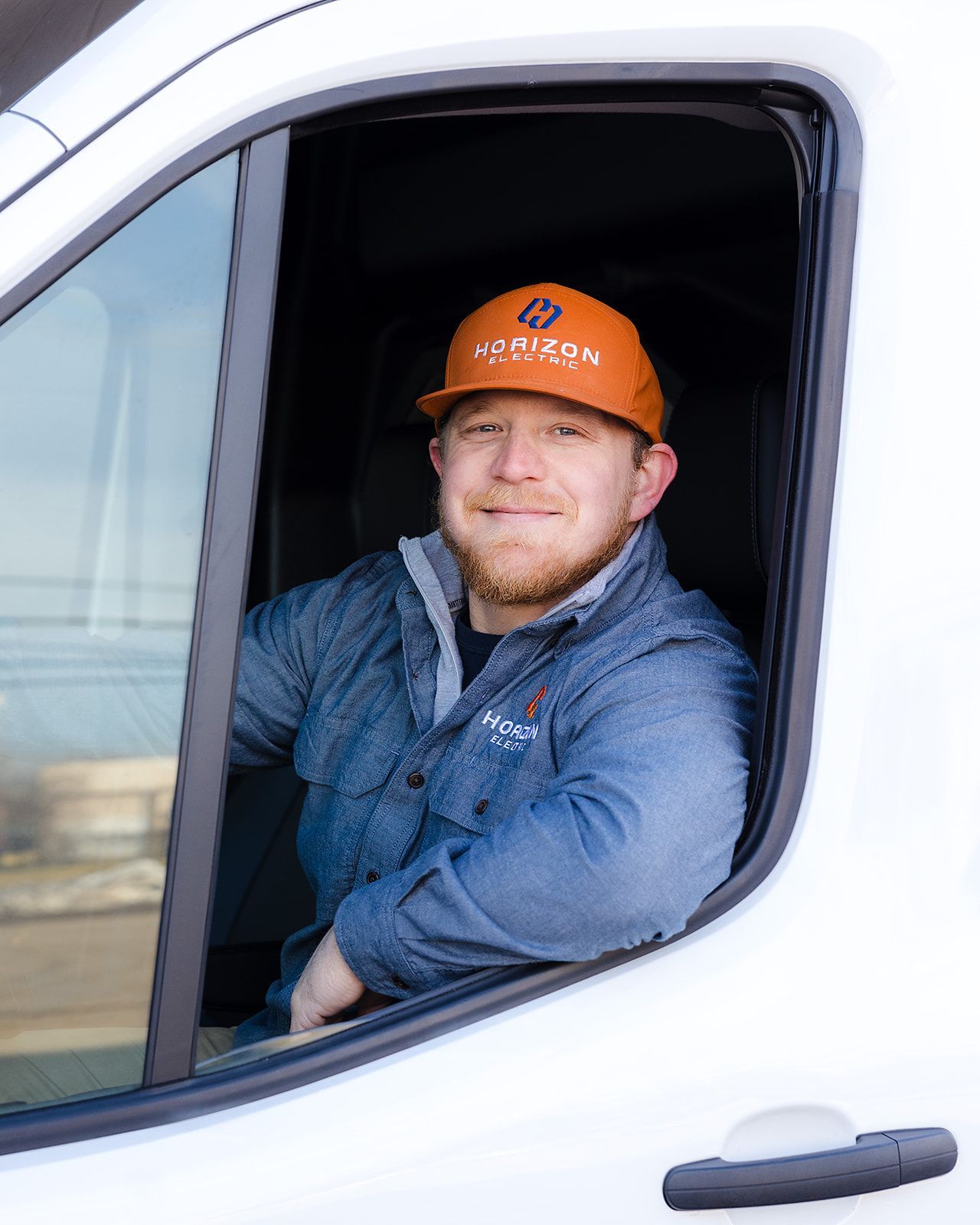 A person wearing a branded orange cap and blue shirt sits in the driver's seat of a white van, smiling at the camera.