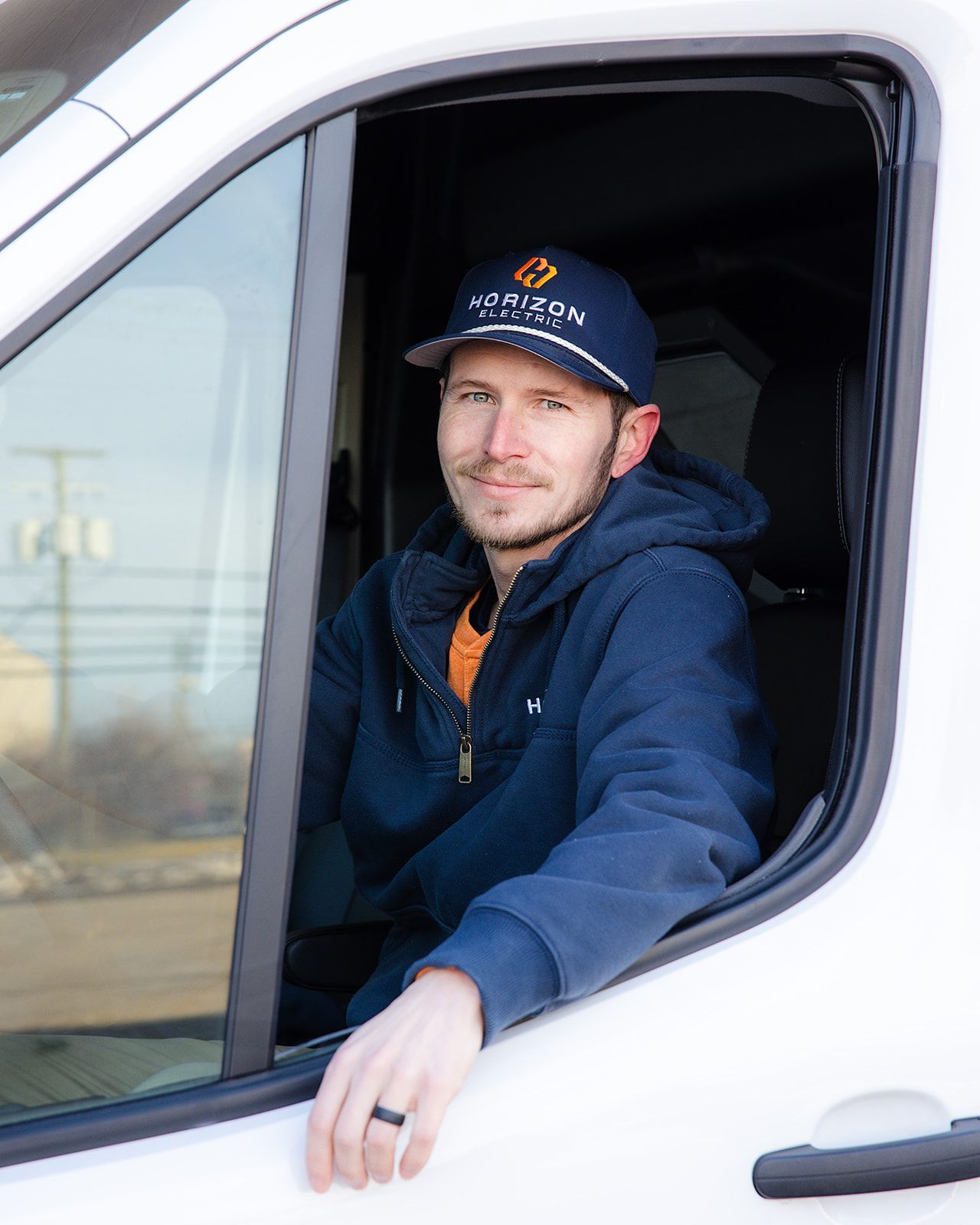 A person in a blue hoodie and branded baseball cap sits in the driver's seat of a white van, looking out the window.