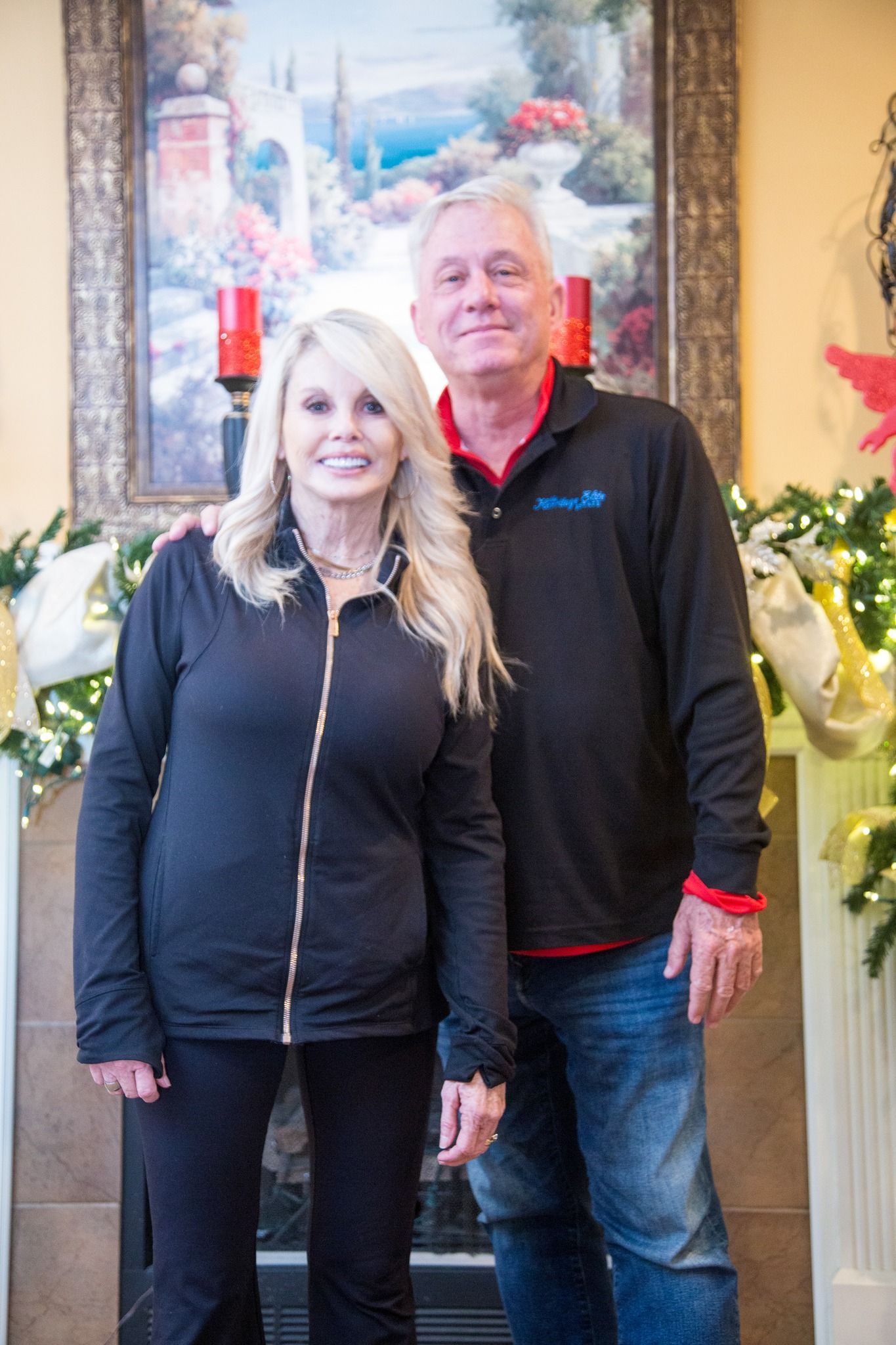 A man and woman in black tops pose together in front of a fireplace with holiday decorations and a framed painting.