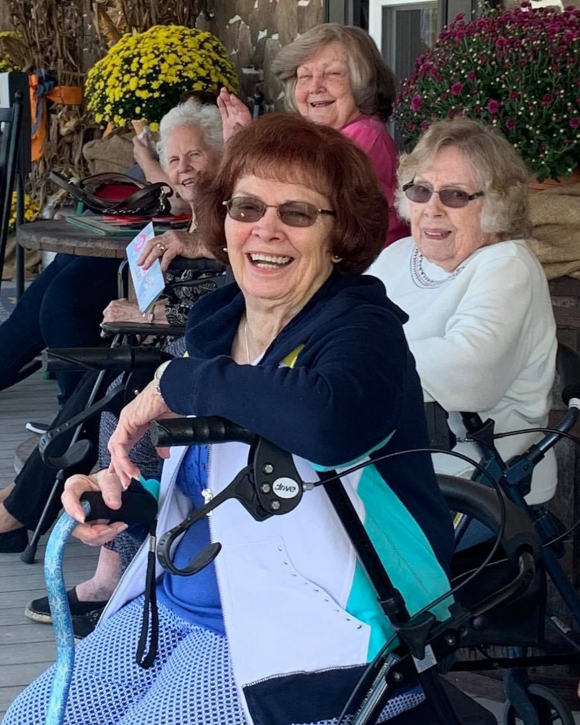 Four people smile outdoors, sitting with walkers and canes in front of pots of yellow and purple flowers near Jacksonville, NC.