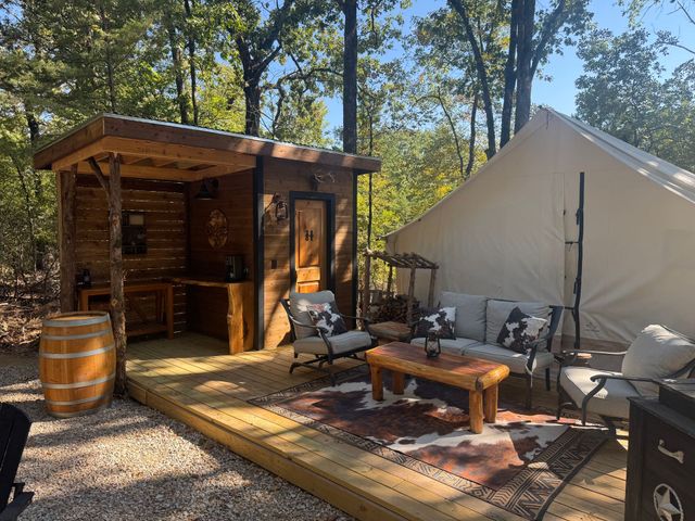 Outdoor campsite with cream-colored tent, wooden platform, and black chairs around a fire pit.