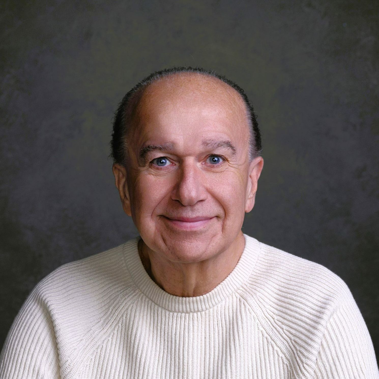 Studio portrait of a smiling man in a cream sweater against a gray background