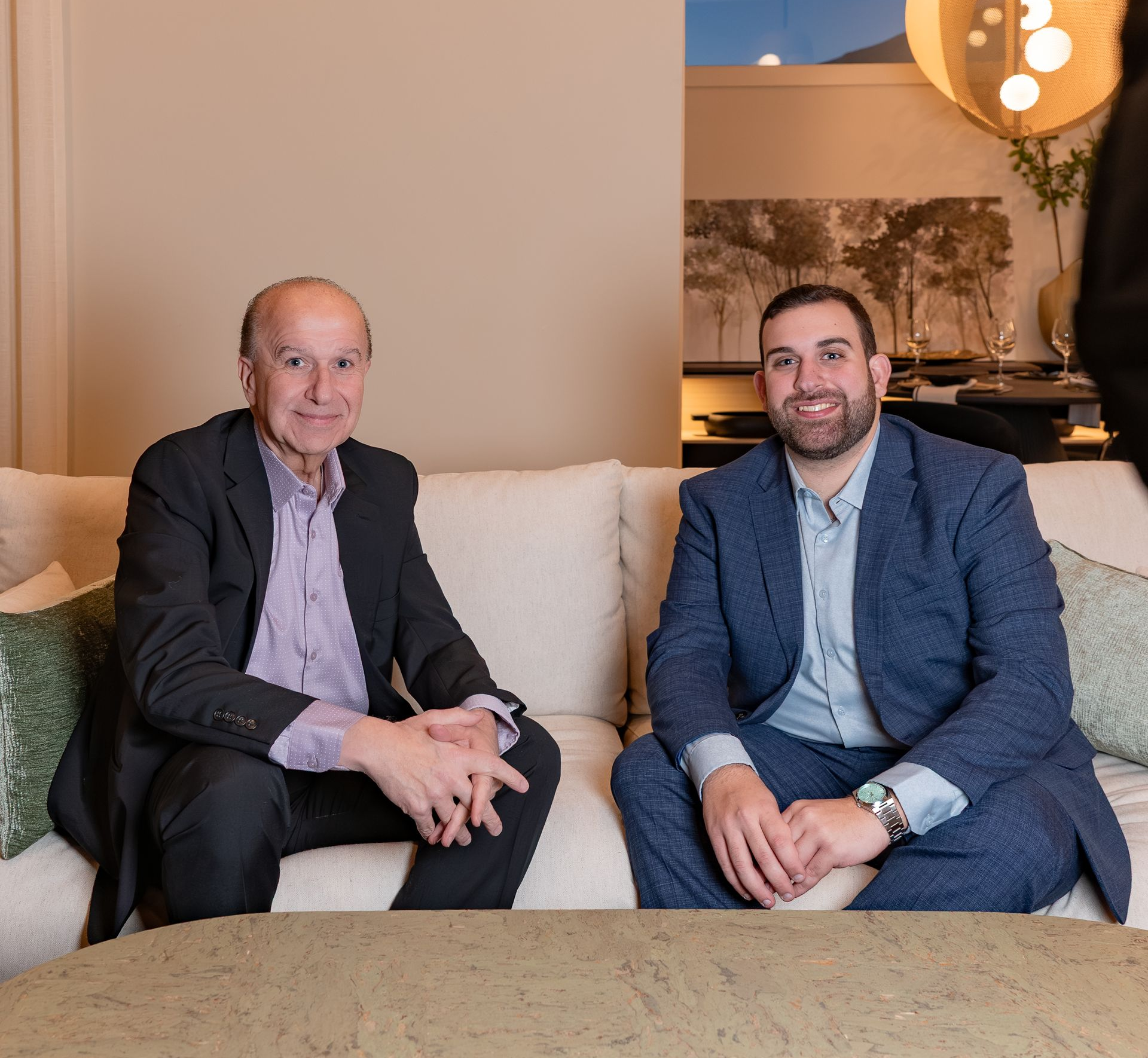 Two men in suits seated on a beige couch in a lounge, smiling at the camera.