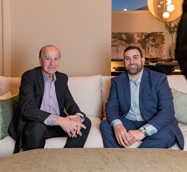 Two men in suits seated on a beige couch in a lounge, smiling at the camera.