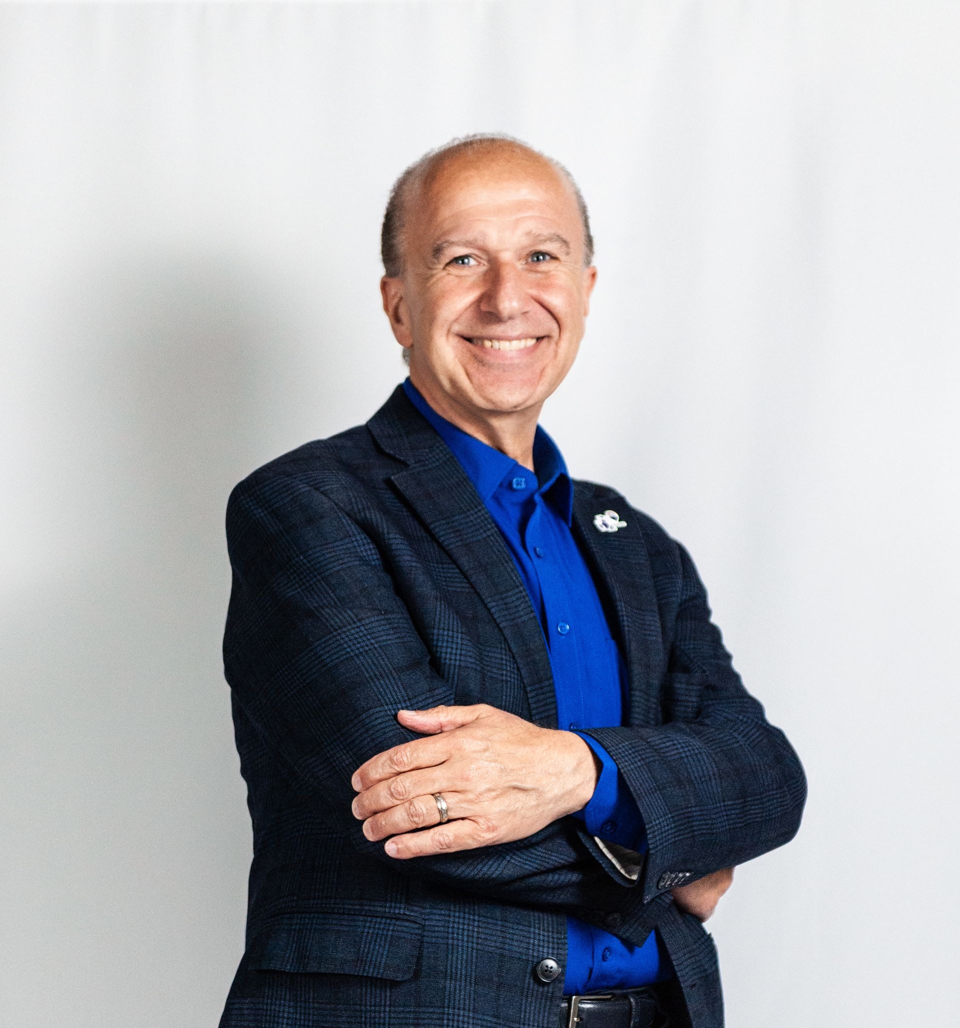 Smiling man in blue shirt and dark blazer, standing with arms crossed against a light background