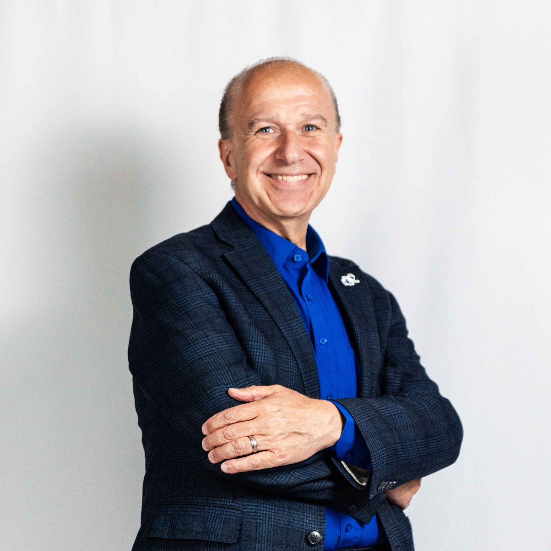 Smiling person in a blue shirt and dark blazer, standing with arms crossed against a light background