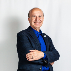 Smiling person in a blue shirt and dark blazer, standing with arms crossed against a light background