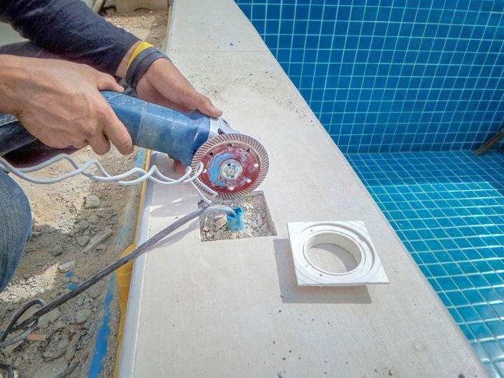Hands holding a blue angle grinder cutting concrete beside a swimming pool drain and blue tile.