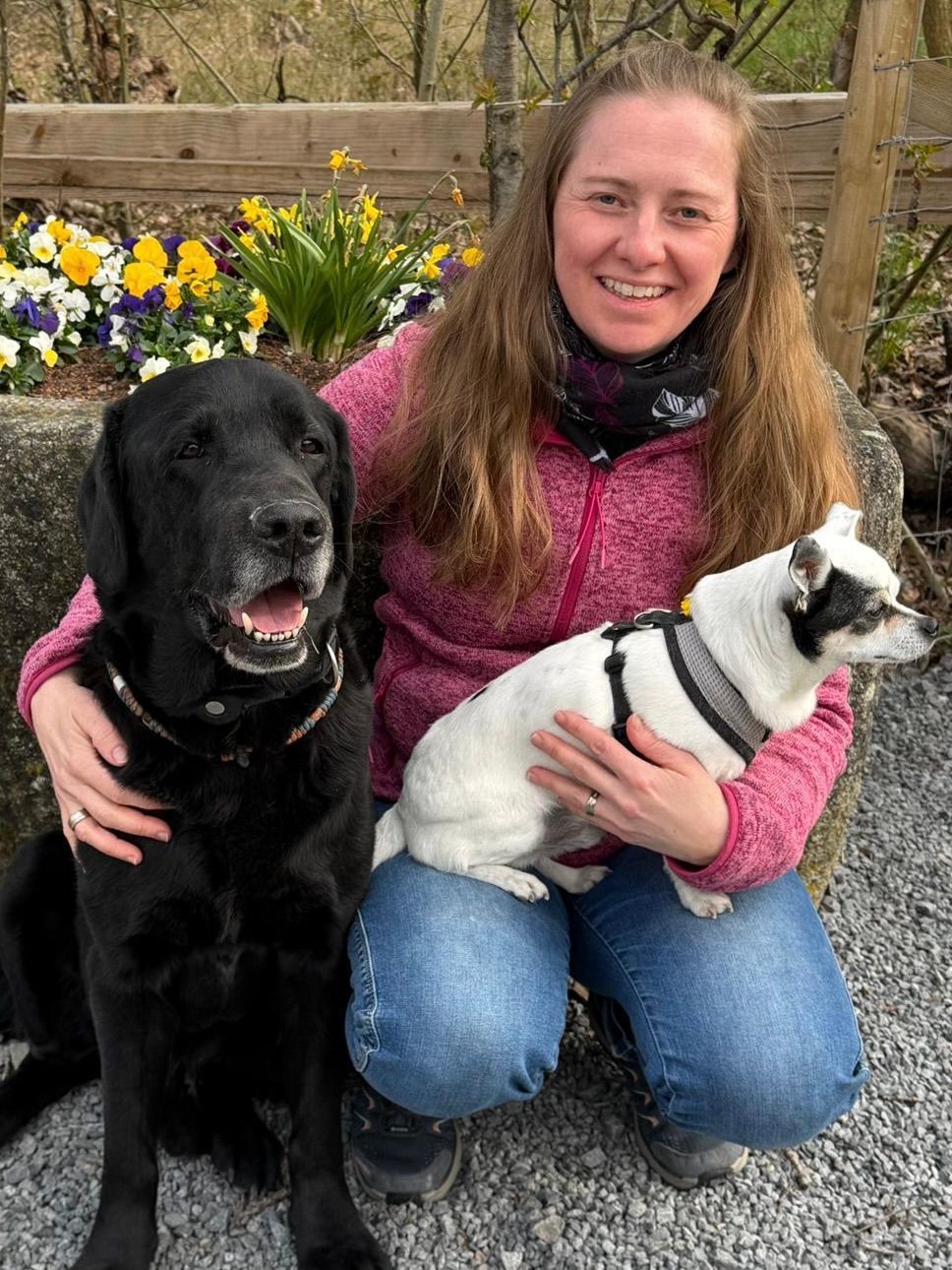 Eine Frau kniet im Freien mit einem schwarzen Hund und einer weißen Katze und lächelt neben Blumen und einem Holzzaun.