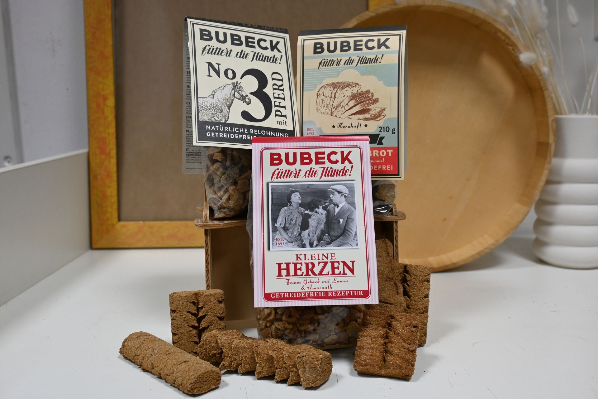 Drei Bubeck-Hundeleckerlidosen mit braunen Keksstäbchen und einem Holztablett auf einer weißen Oberfläche