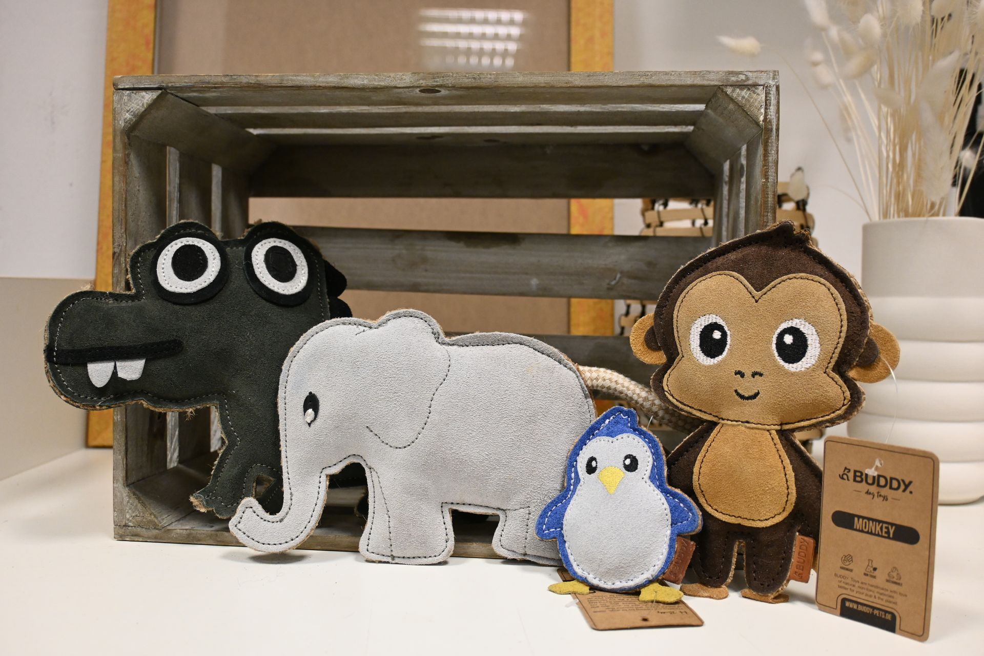 Plüsch-Alligator, Elefant, Pinguin und Affe auf einer Kiste, daneben ein kleines Pappschild.