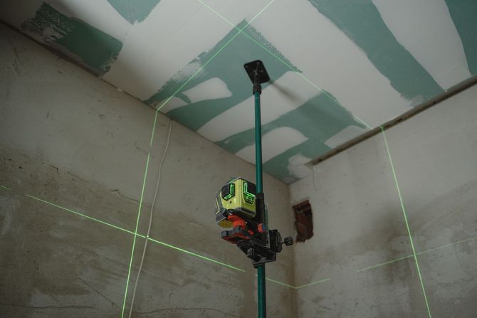 Hand holding a power drill beside a marked measuring strip, with two blurred workers in the background