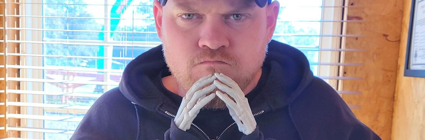 Person in a dark hoodie and cap, looking at the camera with hands clasped under their chin indoors.