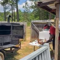 Person in a red shirt working at a table outdoors beside a grill and porch railing.