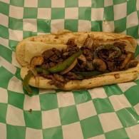 Sandwich with grilled onions and green peppers on green-and-white checkered paper.