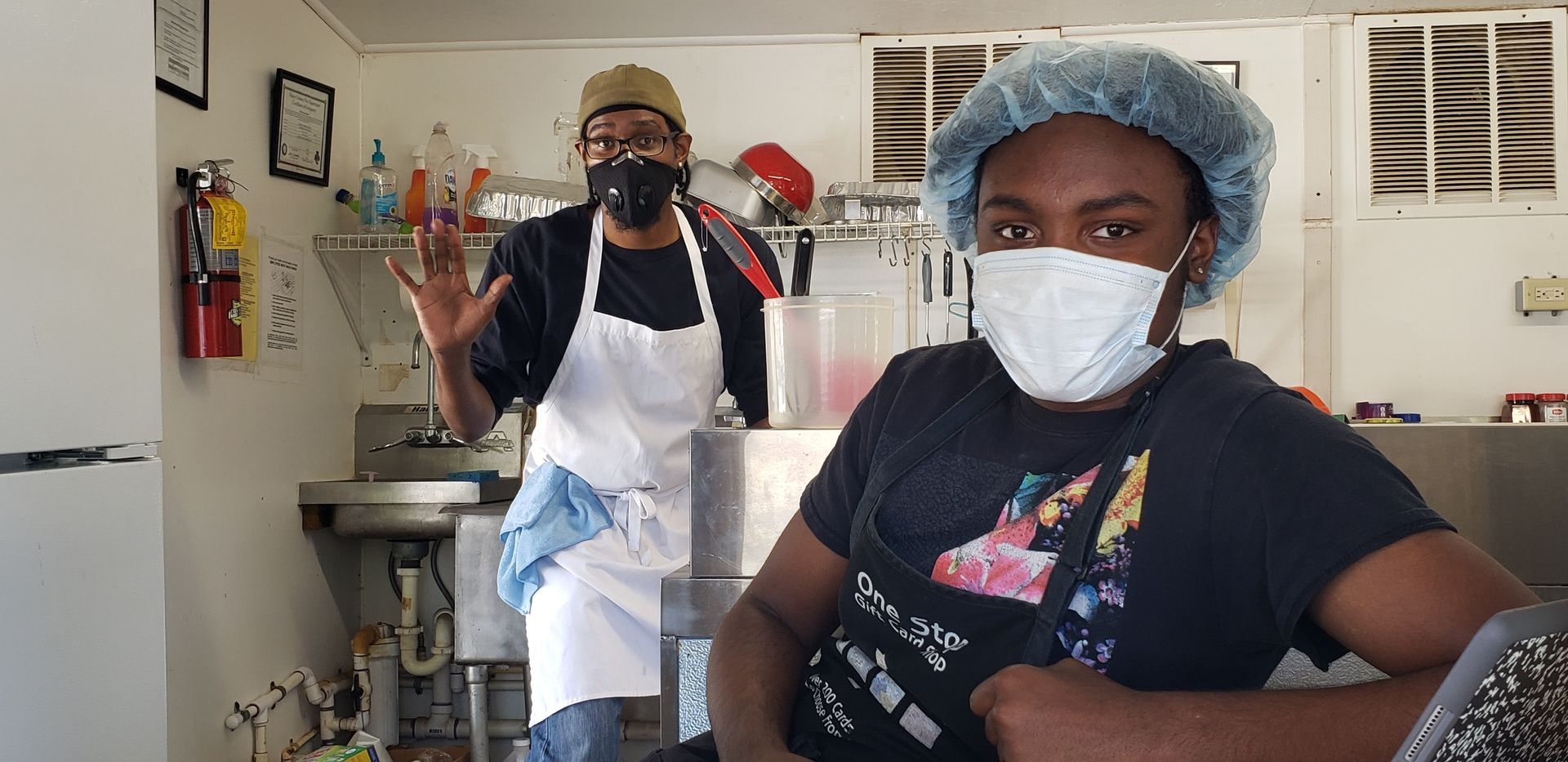 Two masked people in aprons in a kitchen, one seated and one standing and waving.