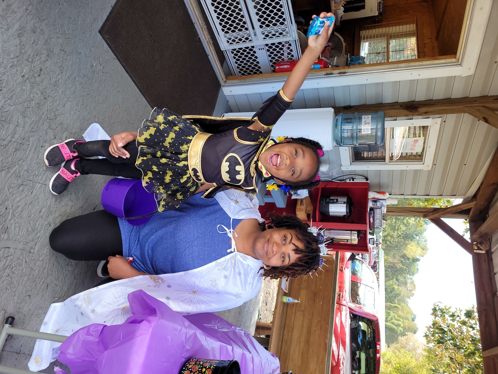 Two people posing on a porch, one holding up a blue item, with purple decorations nearby