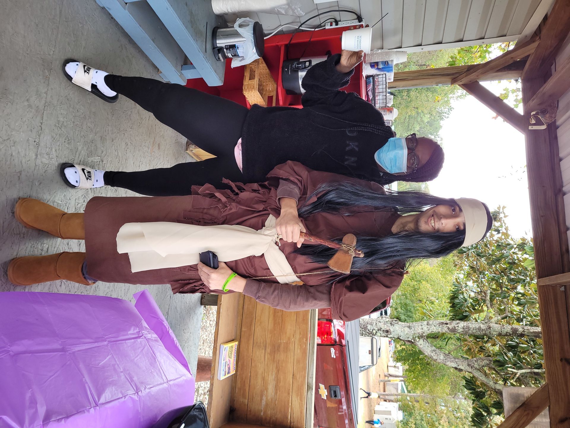 Two people standing outdoors on a porch, wearing masks and winter coats, with shopping bags nearby