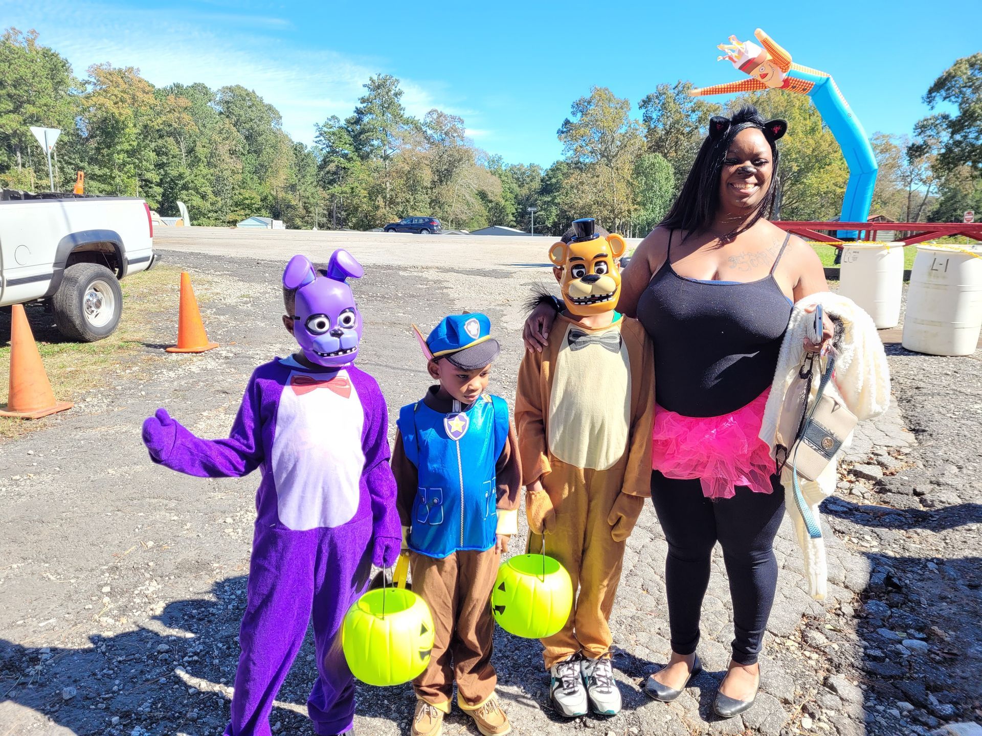 Four people in costumes pose outdoors with Halloween buckets on a sunny gravel lot.
