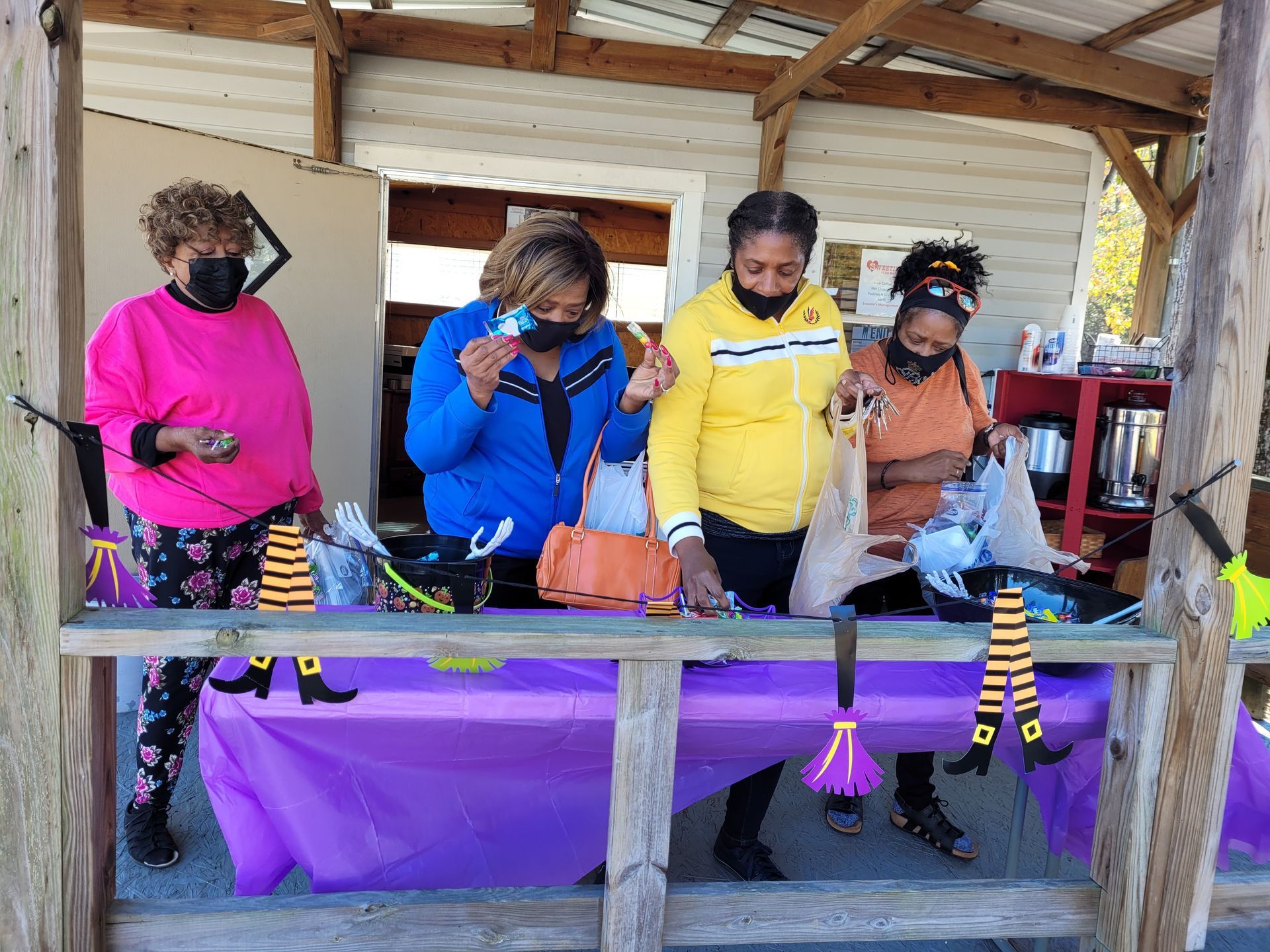 Four masked shoppers examine items at an outdoor craft stall with purple tablecloth and striped stocking decorations