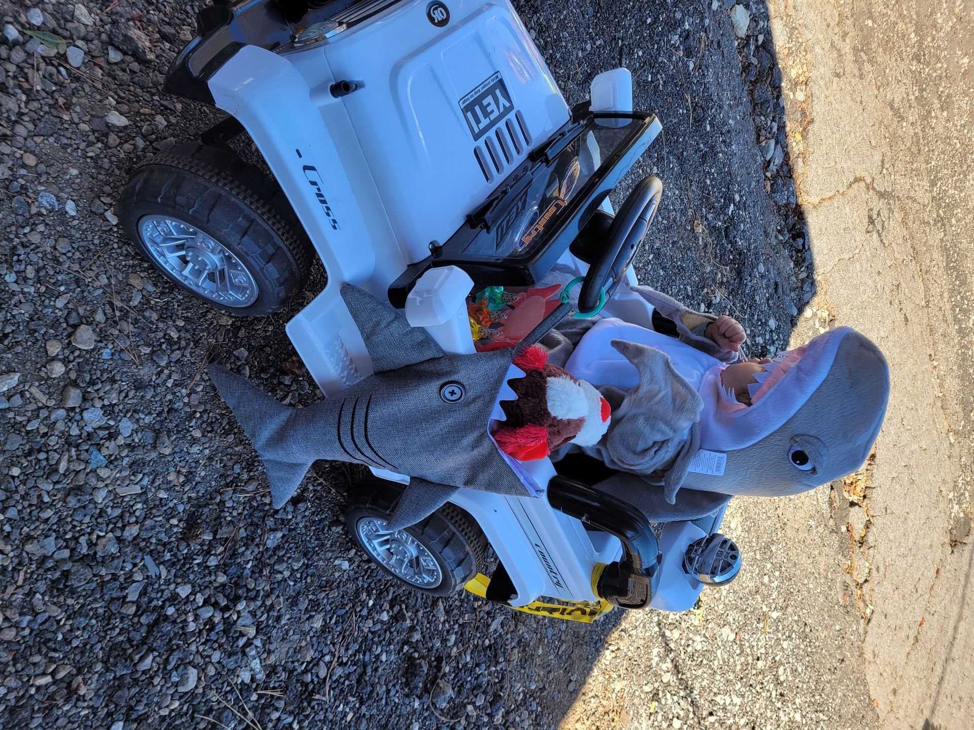 Child in a toy ride-on vehicle outdoors on gravel, holding a red toy and wearing a gray helmet.