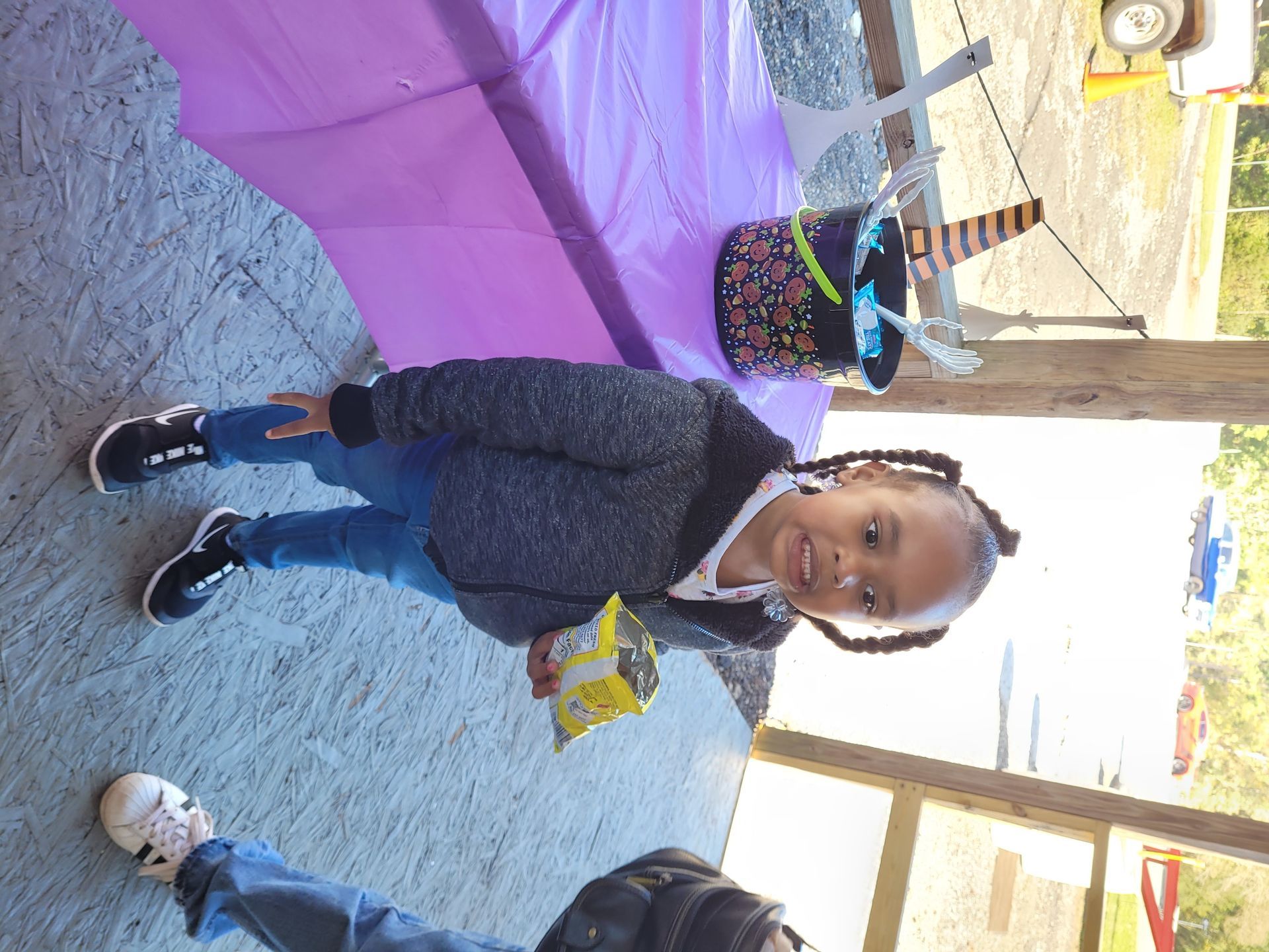 Child standing on a sidewalk beside a purple tent, holding a snack and smiling at the camera.