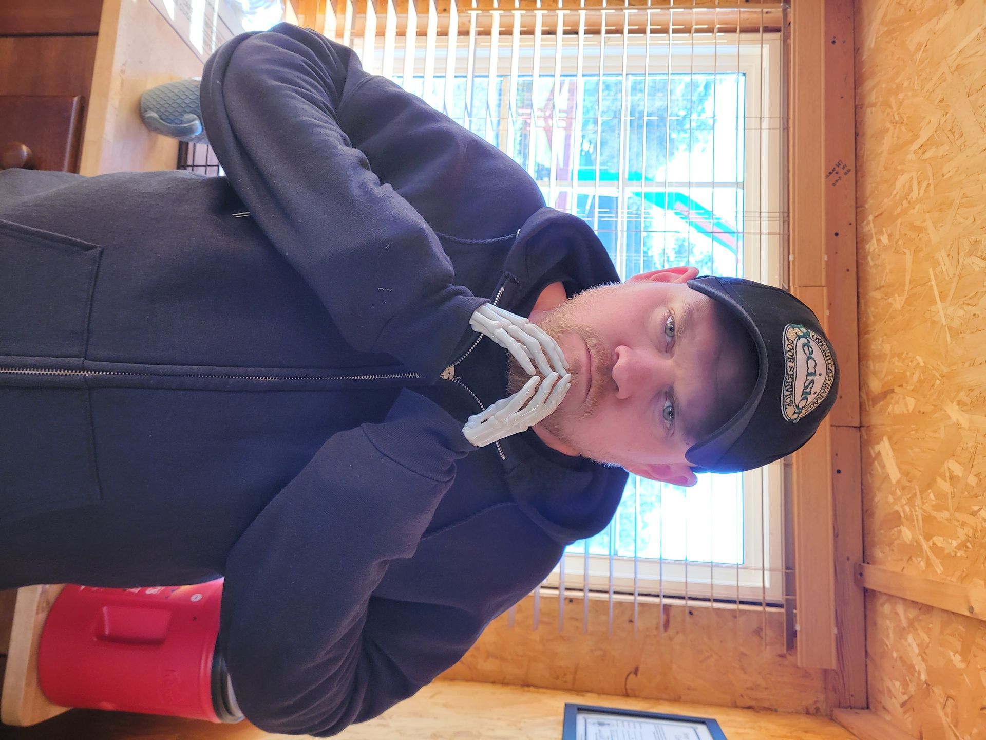 Person in a black hoodie and cap sits indoors by a window, holding a red mug.