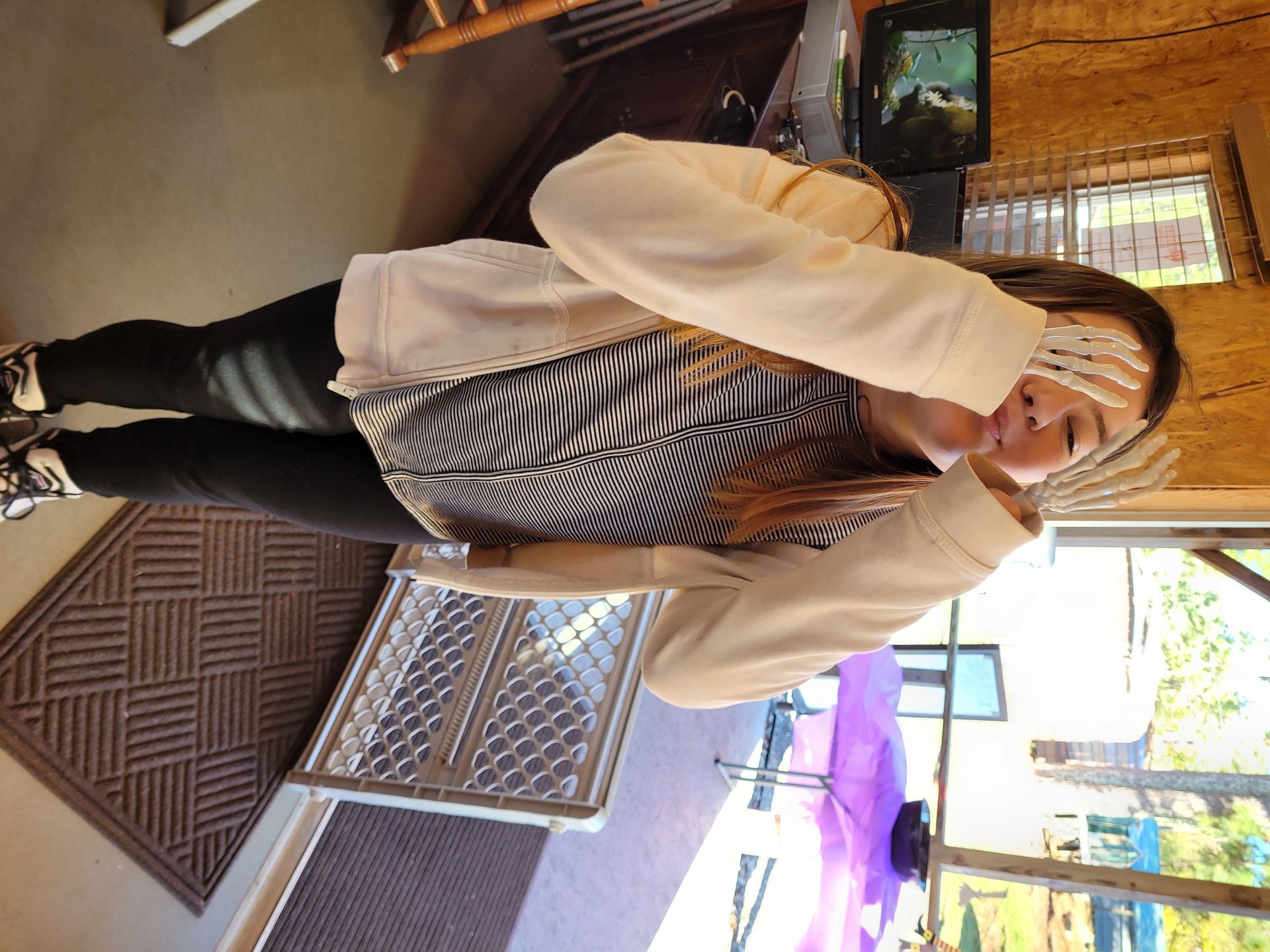 Person indoors, smiling and making a heart gesture, wearing a beige cardigan and striped shirt.