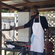 Person grilling food outdoors under a covered patio, wearing a hat, mask, and white apron