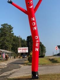 Red inflatable tube man by a roadside shop, with a “NOW OPEN” sign.