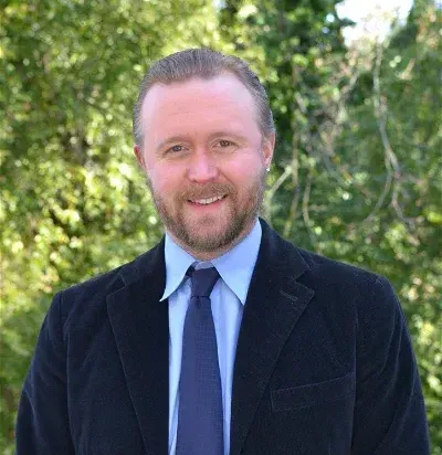 Damon in a dark suit with a blue tie, smiling, against a blurred green background.
