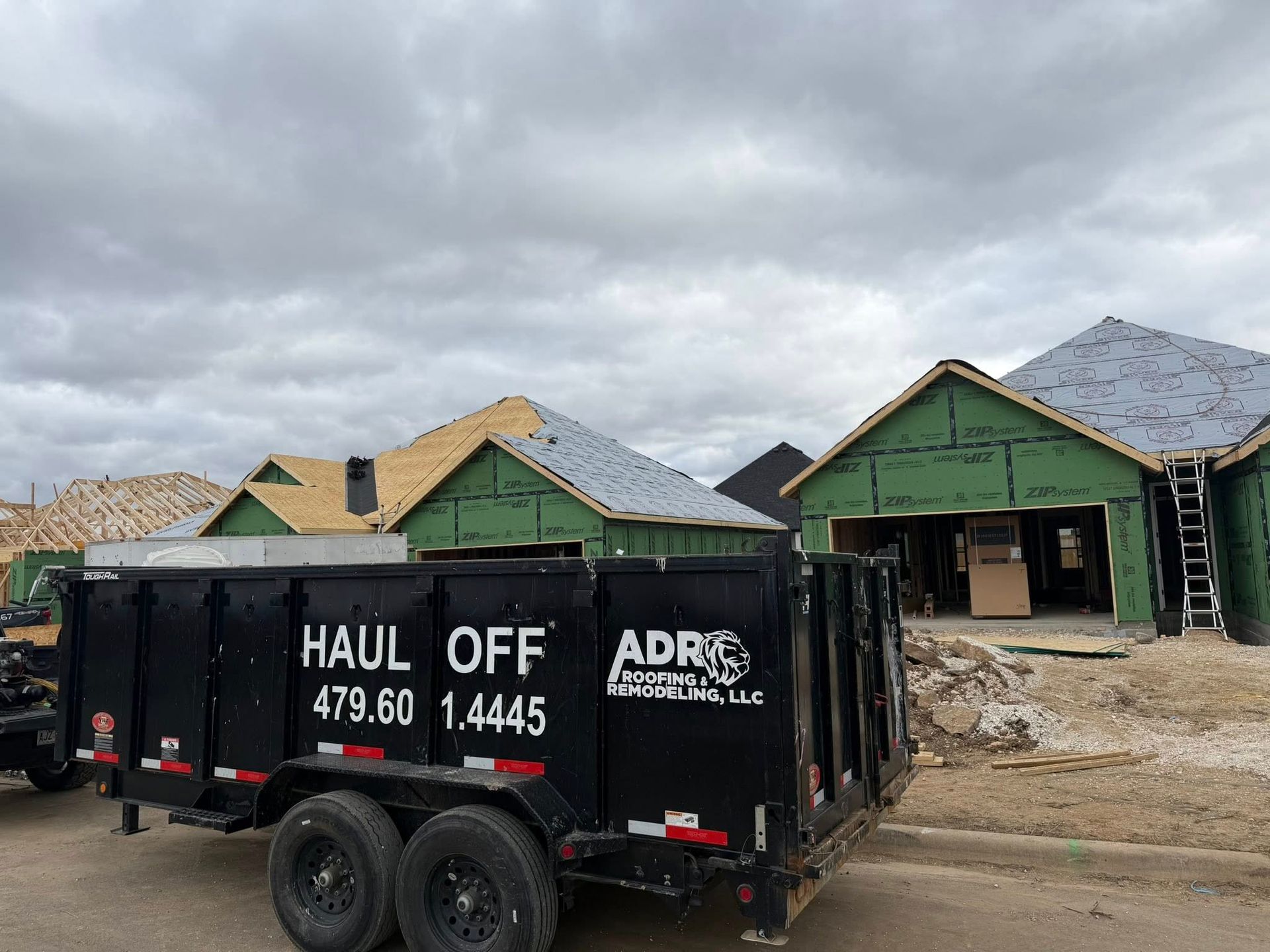 A black ADR Haul Off dumpster trailer sits in front of two houses under construction with exposed wooden framing.