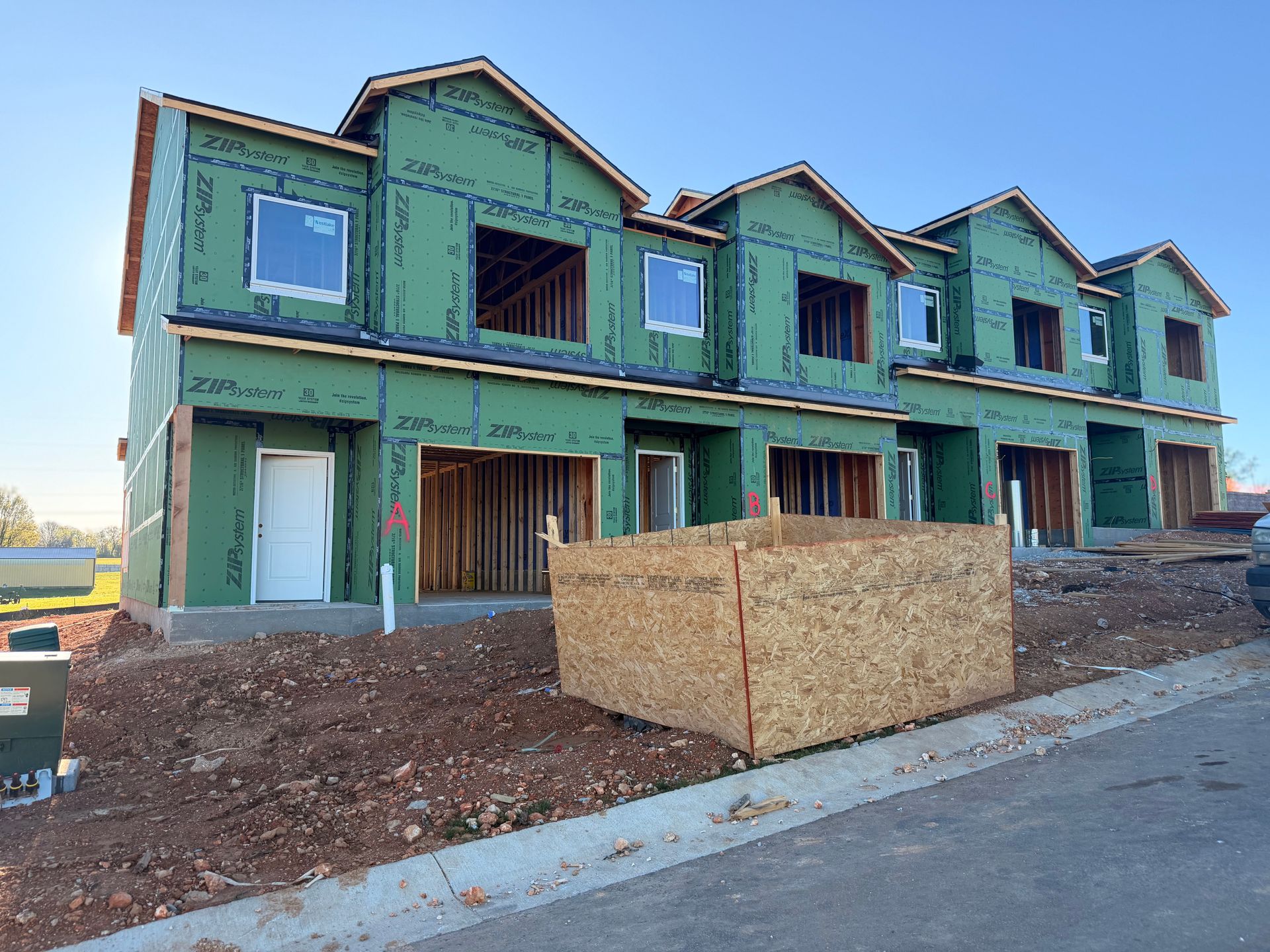 A row of multi-story townhouses under construction, wrapped in green protective siding, with a wooden crate in front.