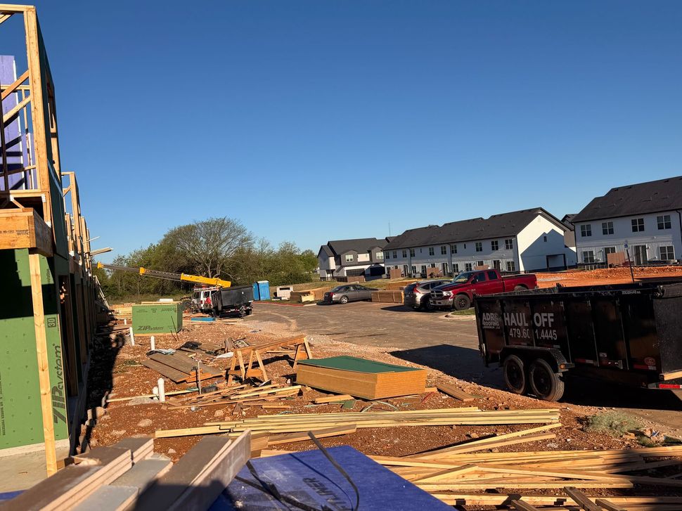 Construction site with unfinished building framing, materials, and a dumpster in a residential neighborhood under a sky.
