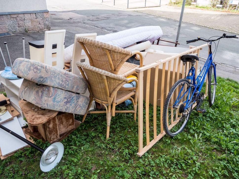 A pile of discarded items, including a blue bicycle, wicker chairs, cushions, and a wooden gate, placed on a grassy curb.