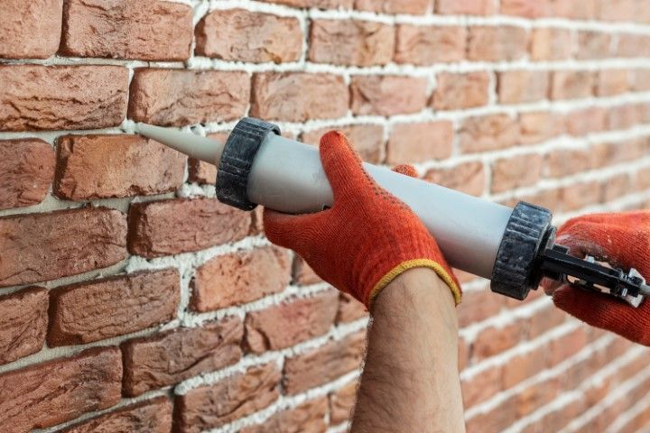 Gloved hands caulk a brick wall crack with a caulking gun