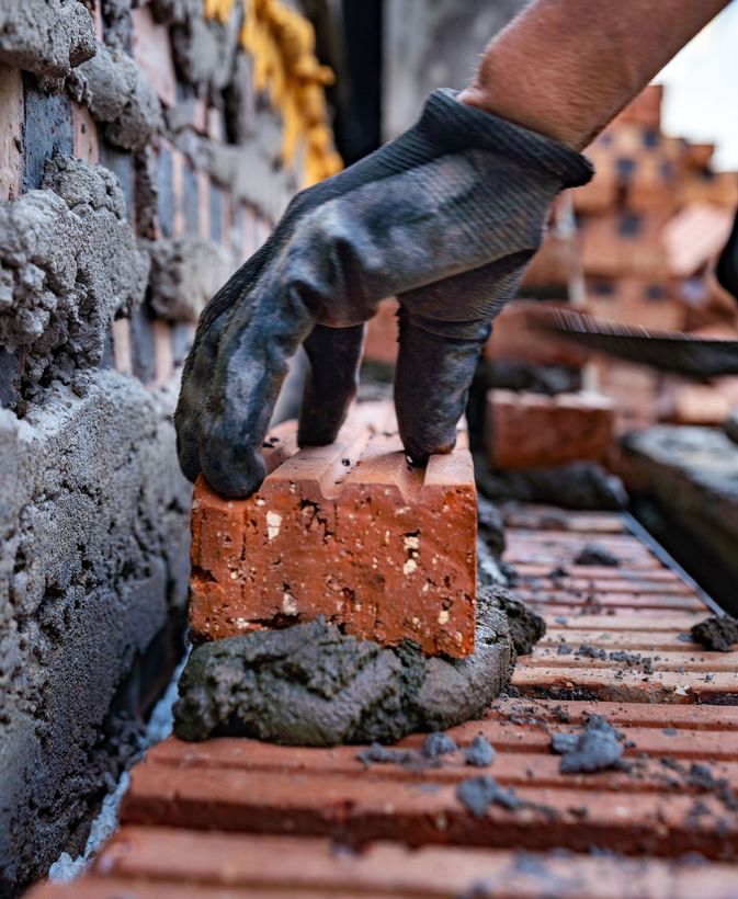 Gloved hand placing a red brick with mortar on a brick wall under construction