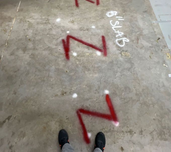 Concrete floor with red spray-painted zigzags and white “SLAB” marking, viewed from above with shoes visible.