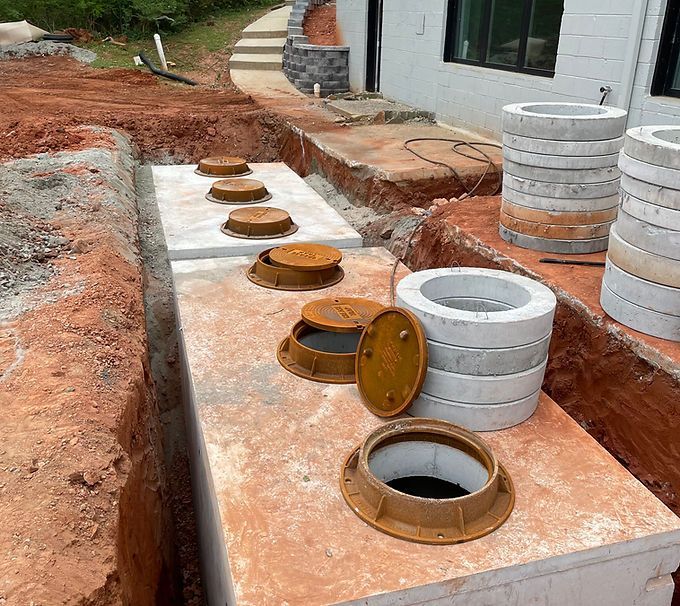 Open septic tank lids and concrete risers in a trench beside a house under construction.