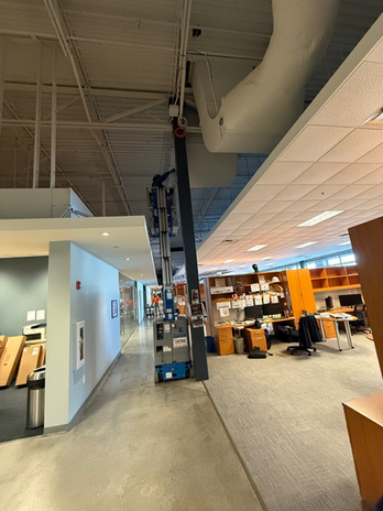 Office interior with a tall ladder on a central column, desks, chairs, and orange-lit workstations.