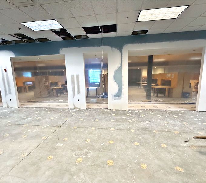 Empty office with glass partitions, damaged ceiling tiles, and debris-stained concrete floor.