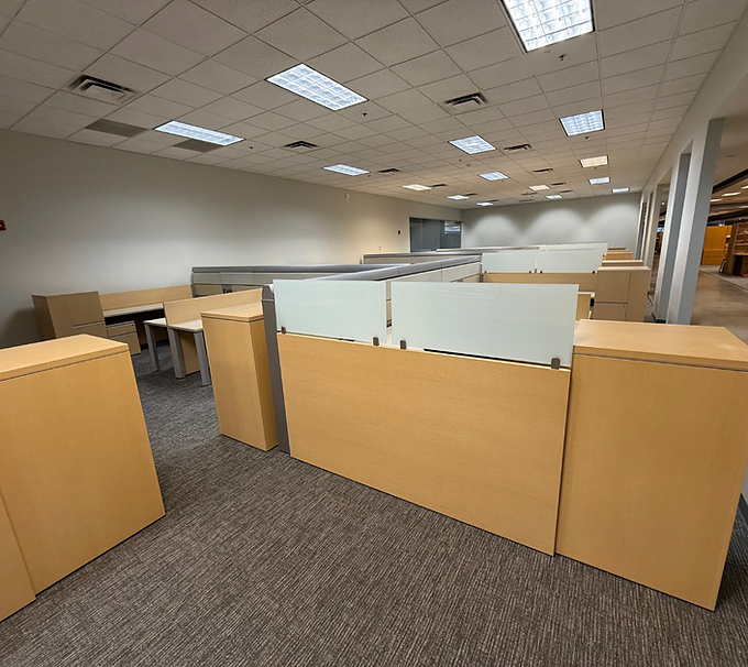 Empty office cubicles with tan partitions and carpeted floor under fluorescent ceiling lights.