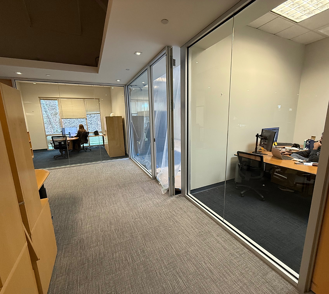 Office hallway with glass walls, leading to desks and seated coworkers in a quiet workspace.