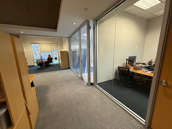 Office hallway with glass-walled rooms, carpeted floor, and desks visible inside.