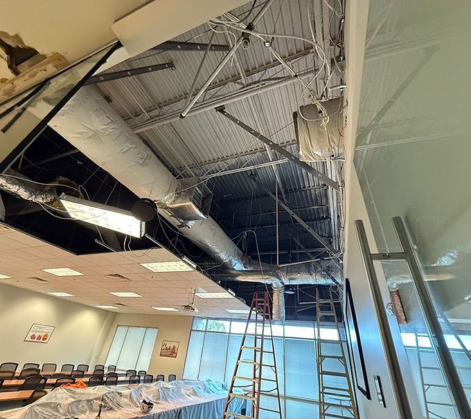 Damaged office ceiling with exposed ducts, a ladder, and debris in a room with chairs and tables